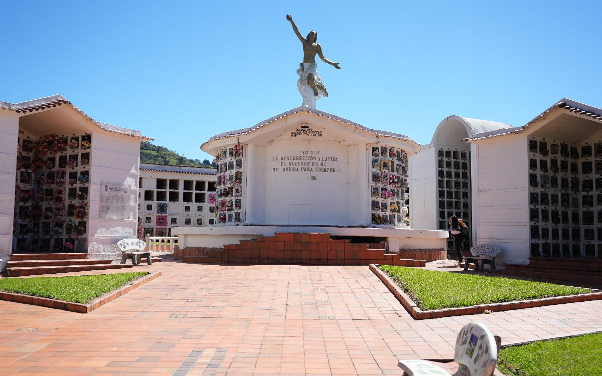 Fotografía a plena luz del día del patio principal del cementerio. En el centro destaca un monumento de pared blanca con la inscripción «Yo soy la resurrección y la vida», coronado por una estatua de Jesucristo con los brazos abiertos. El monumento está flanqueado por estructuras con nichos funerarios y rodeado por un piso cerámico con jardineras triangulares y bancas de piedra.