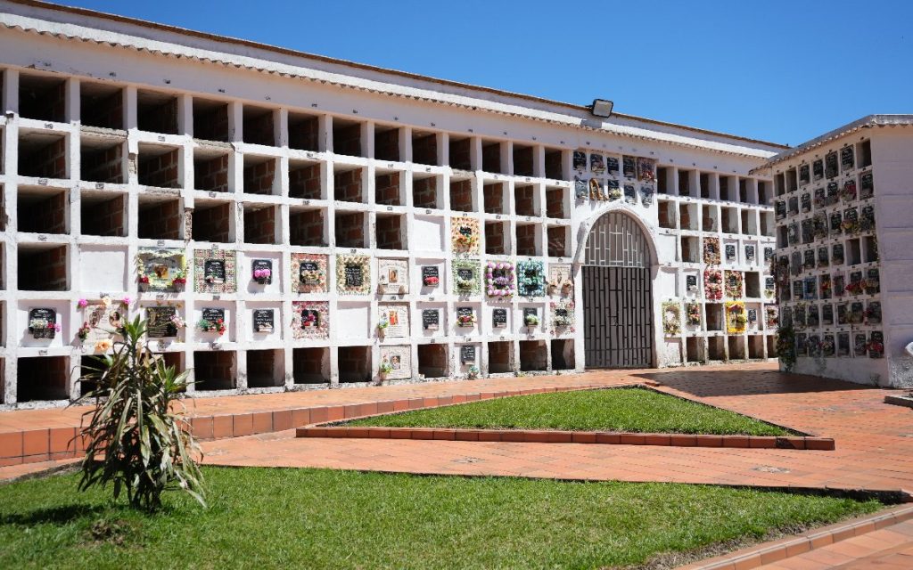Fachada rectilínea de una estructura funeraria de dos a tres niveles con cuadrículas de nichos sobre ladrillo expuesto y blanco. En el centro hay una entrada con arco de medio punto y portón de reja metálica. Muchas de las bóvedas inferiores tienen lápidas decoradas con flores y placas. En primer plano hay senderos de baldosas rojas y jardineras con pasto verde.