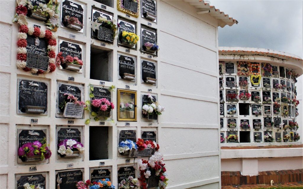 Toma en plano medio de la esquina de un edificio blanco del cementerio. La pared frontal muestra nichos organizados en retícula; varias lápidas de mármol negro están adornadas con abundantes coronas de flores artificiales en tonos rojos, blancos y amarillos, además de fotografías y recuerdos. Al fondo se vislumbra otra estructura circular de bóvedas bajo un cielo nublado.