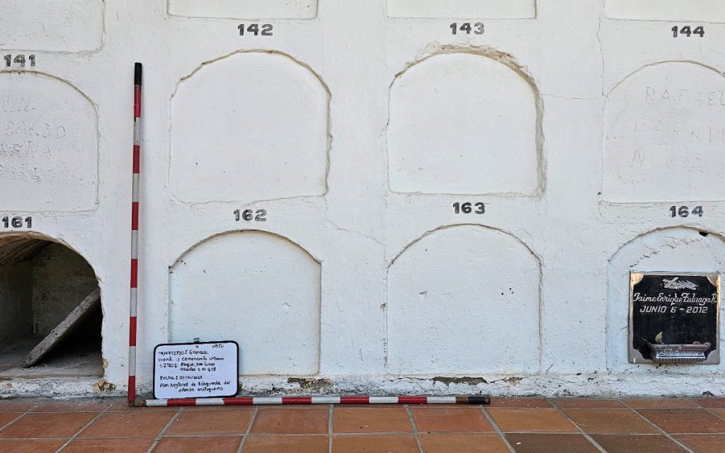Plano detalle de una pared blanca con arcos marcados para bóvedas con números impresos (141, 142, 162, 163, entre otros). En la base se observa una regla de escala topográfica o forense de franjas rojas y blancas en forma de L junto a una pequeña pizarra de identificación del sitio. A la derecha, una de las bóvedas conserva una placa de mármol negro fechada en junio de 2012.