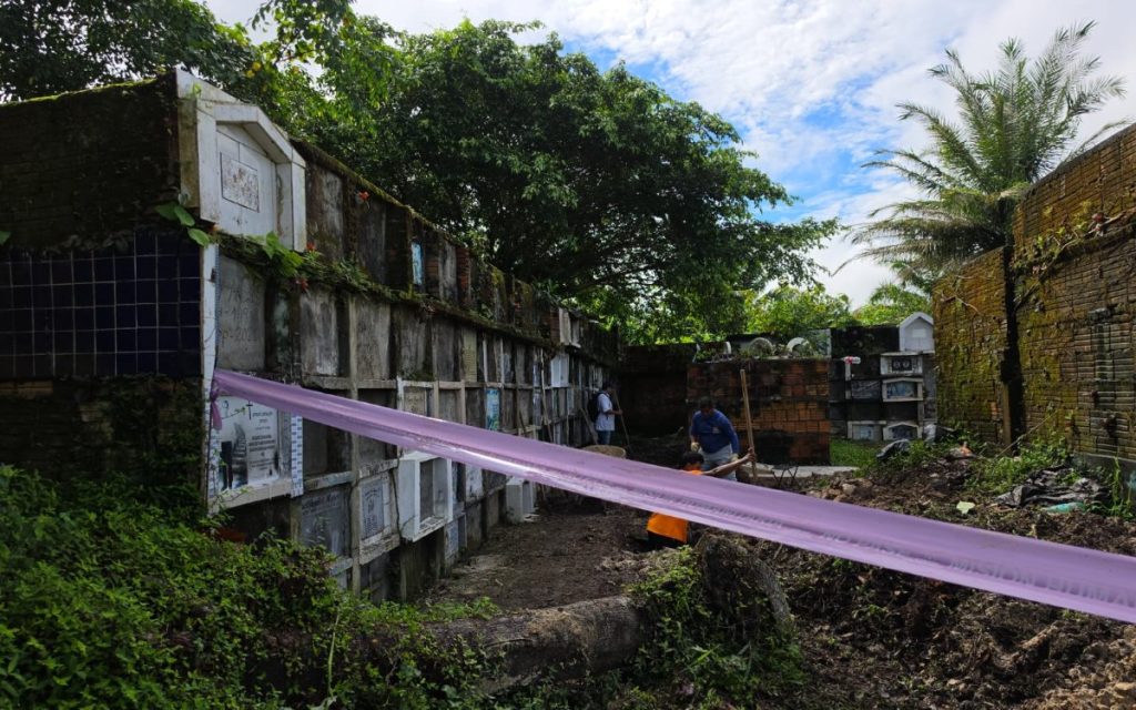 Toma en perspectiva del pasillo de un cementerio antiguo. Una cinta morada atraviesa la escena en primer plano delimitando el área. Al fondo, personal con palas trabaja cerca de las bóvedas en un entorno rodeado de vegetación y árboles.