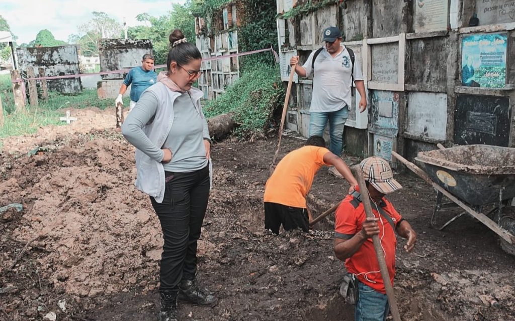 Plano medio de un equipo de trabajo en el cementerio. En primer plano, una mujer vestida con chaleco claro y gafas observa atentamente mientras operarios con palas excavando en la tierra húmeda cerca del muro de nichos.