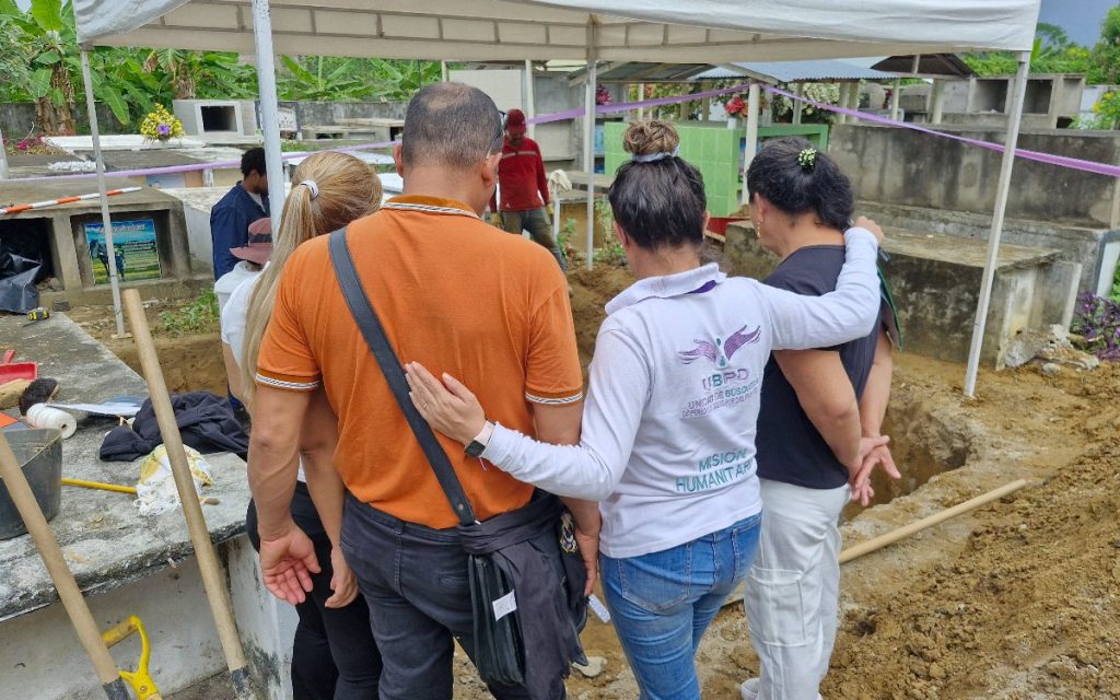 Un grupo de personas abraza solidariamente a un hombre y una mujer frente a una fosa abierta en un cementerio. Una integrante de la Unidad de Búsqueda de Personas dadas por Desaparecidas (UBPD), vistiendo un buzo con la inscripción "Misión Humanitaria", los acompaña de espaldas bajo una carpa blanca.