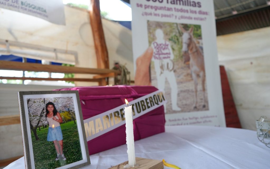 Mesa ceremonial cubierta con un mantel blanco donde reposa un cofre fúnebre rosado con el nombre "MARIBEL TUBERQUIA" en cinta dorada, una vela encendida, el retrato de una niña y un pasacalle de fondo de la Unidad de Búsqueda de Personas dadas por Desaparecidas.