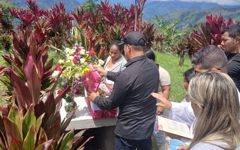 Familiares y acompañantes reunidos al aire libre en un cementerio rodeado de vegetación y montañas. Un hombre coloca un cofre rosado cubierto de flores en una bóveda, mientras una mujer a su lado sostiene un libro abierto durante una ceremonia de inhumación.