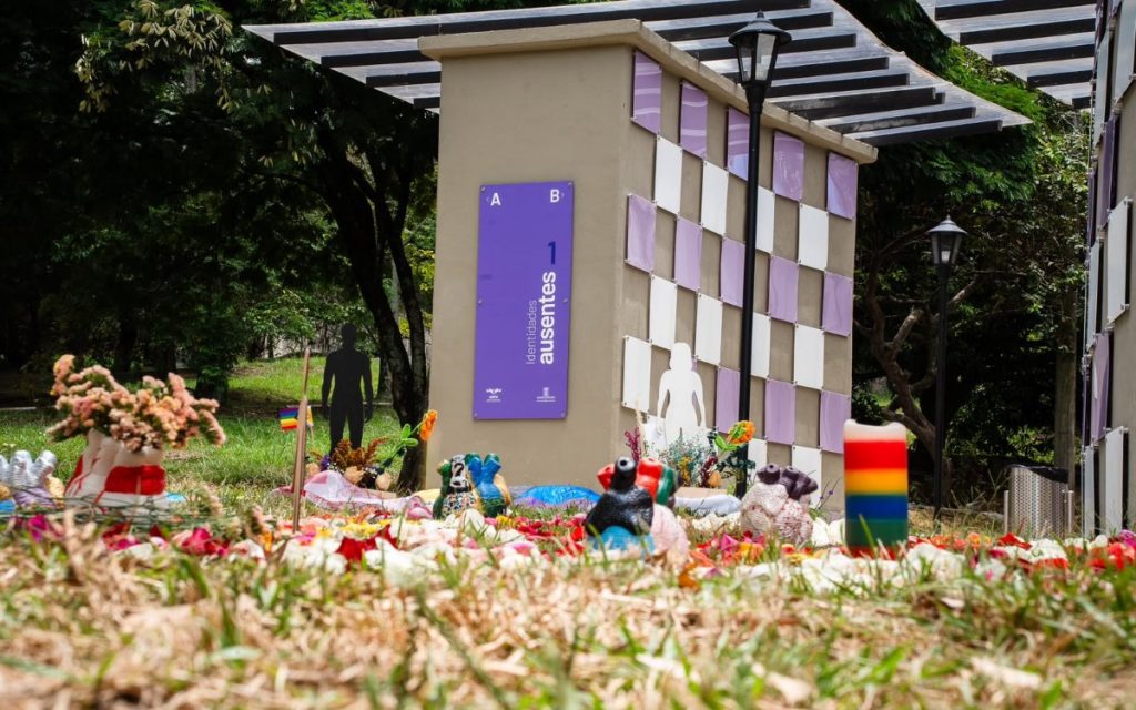 Fotografía en plano general medio al aire libre de un área conmemorativa. En el primer plano, el suelo de césped está cubierto de pétalos de flores de diversos colores, cerámicas artesanales en forma de corazones o torsos y un velón con franjas con los colores de la bandera LGBTIQ+. De fondo, se aprecia un módulo arquitectónico de nichos con un patrón tipo ajedrez en tonos lila y blanco. Sobre el muro gris principal hay un cartel morado con el texto vertical «Identidades ausentes 1». A los lados hay siluetas negras y blancas de figuras humanas y vegetación al fondo.