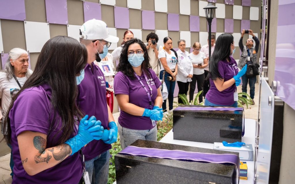 Fotografía en plano medio de un grupo de personas durante una ceremonia o acto conmemorativo. En primer plano, miembros del equipo técnico de la Unidad de Búsqueda de Personas dadas por Desaparecidas (UBPD) llevan camisetas moradas institucionales, tapabocas y guantes de nitrilo azules; observan con actitud respetuosa dos cofres u osarios de madera sobre una mesa cubierta, decorados con cintas de color lila. Al fondo, familiares y asistentes observan atentamente frente a un muro con paneles lila y blanco.