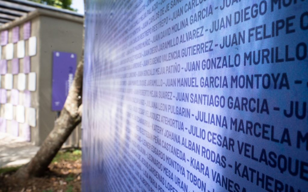 Plano en ángulo oblicuo y de cerca de un mural o lienzo azul con una lista extensa de nombres impresos en mayúsculas sostenidos sobre un fondo textil. Al fondo se aprecia parcialmente la vegetación del parque y estructuras del recinto.