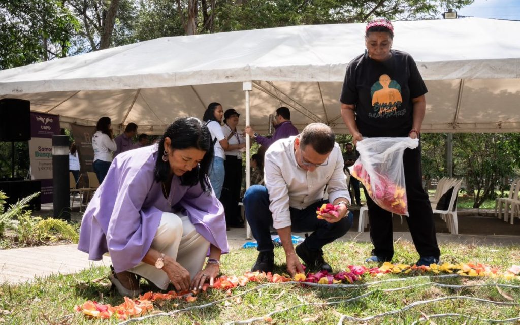 Varias personas agrupadas al aire libre bajo una carpa blanca realizando una ofrenda floral. Dos personas agachadas organizan cuidadosamente un camino o espiral trazado con pétalos de flores amarillas y naranjas sobre el pasto, mientras una mujer de pie sostiene una bolsa con más pétalos.