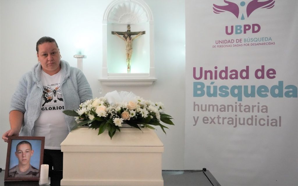 Una mujer con chaqueta azul sosteniendo con la mano un portarretratos con la foto de un joven. Al centro, un pequeño cofre funerario de color blanco con un arreglo de flores blancas sobre él y un crucifijo en la pared de fondo. A la derecha, se observa un pendón con el logo y texto de la "UBPD - Unidad de Búsqueda humanitaria y extrajudicial".