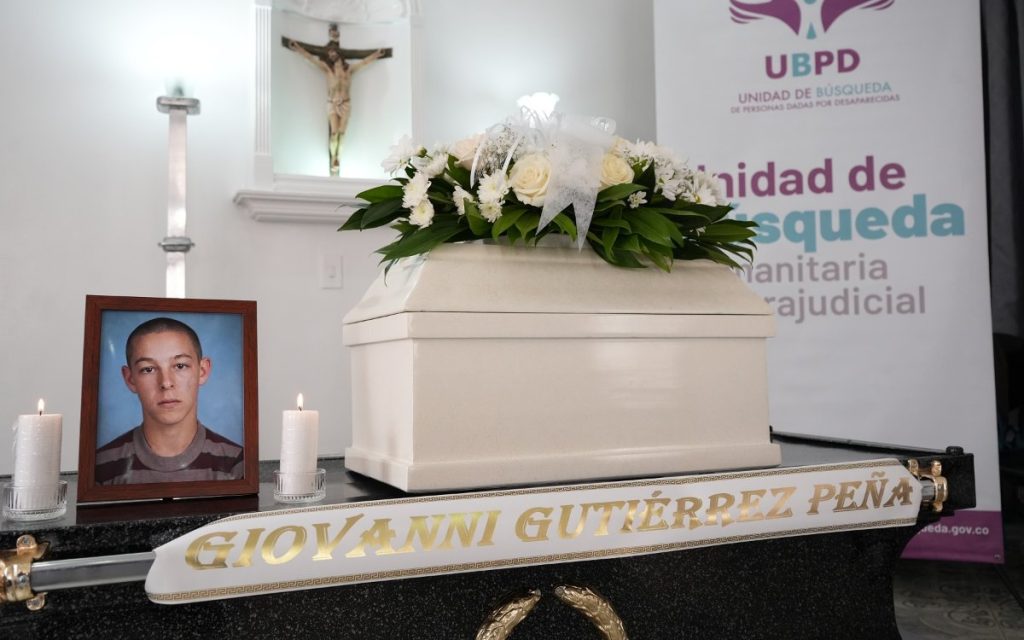 Plano medio del cofre blanco fúnebre cubierto con un arreglo floral. Frente al cofre, a la izquierda, se encuentra el retrato enmarcado del joven Giovanni Gutiérrez flanqueado por dos velas blancas encendidas. En el borde inferior, una cinta blanca lleva impreso en letras doradas el nombre "GIOVANNI GUTIÉRREZ PEÑA". Al fondo se aprecia el pendón de la UBPD.