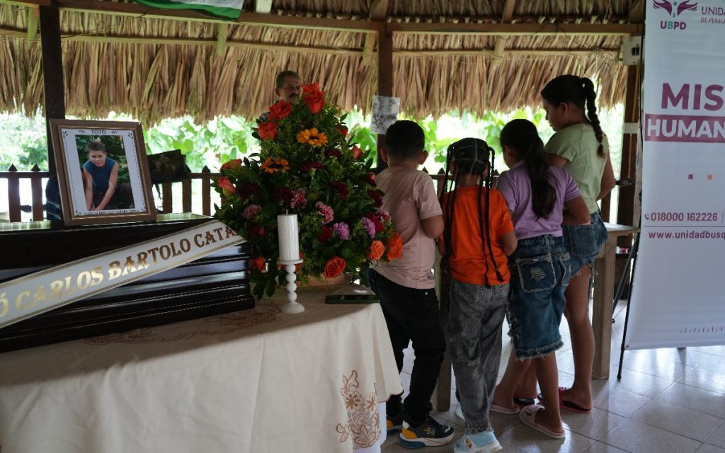 En el interior de un bohío con techo de paja, cuatro niños están de espaldas observando un altar. A la izquierda se encuentra un cofre fúnebre de madera con una cinta blanca que dice "Carlos Bartolo Cataño". Sobre el cofre descansa un retrato con la palabra "Toto". Al lado hay un arreglo floral con rosas, un girasol y una vela blanca encendida. A la derecha se aprecia un pendón de la UBPD con el texto "Misión Humanitaria".