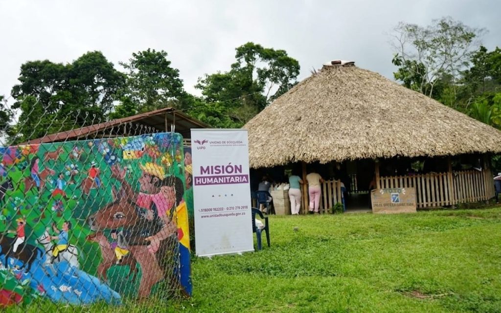 Construcción comunitaria de madera con techo grande de paja cónico rodeada de césped. En primer plano a la izquierda hay una cerca con un mural multicolor que ilustra personas a caballo. Al lado del mural se encuentra un pendón blanco de la Unidad de Búsqueda de Personas dadas por Desaparecidas con el lema "Misión Humanitaria". Al fondo se ven personas reunidas bajo la estructura.