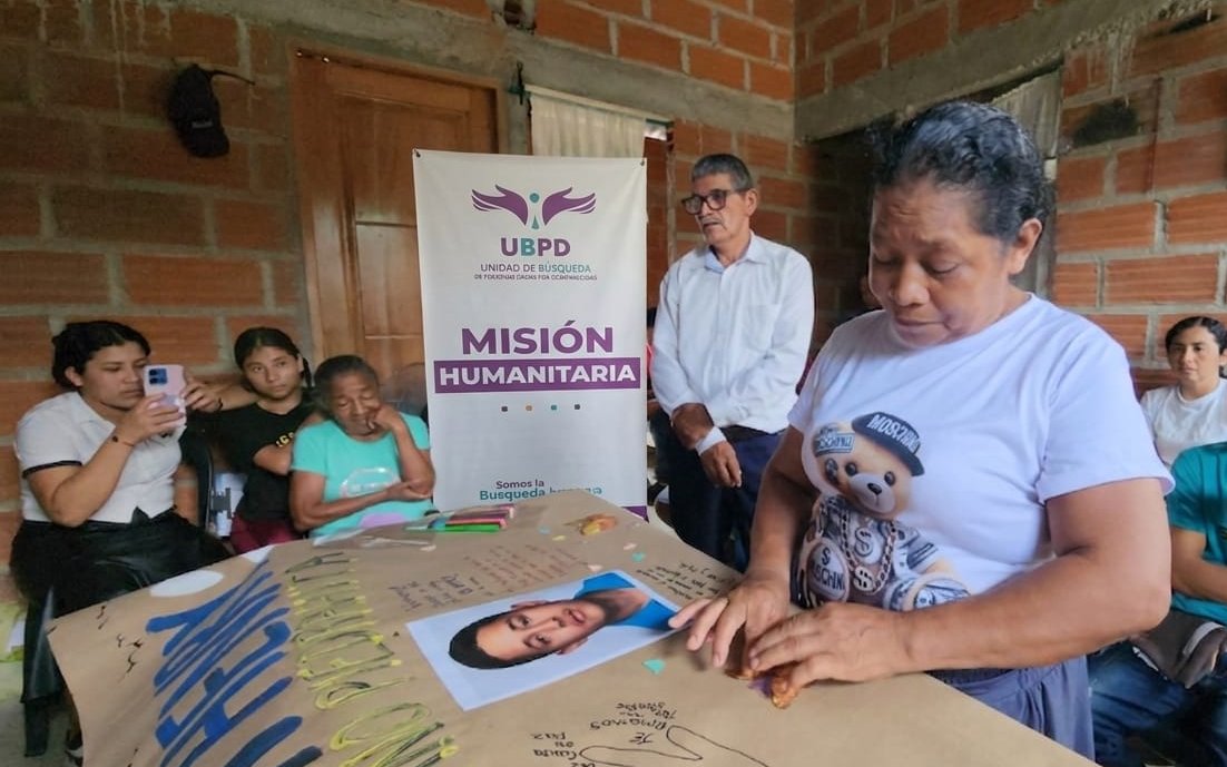 Una mujer con camiseta blanca observa atentamente una fotografía impreso del rostro de Jhon Henry colocada sobre un cartel de papel marrón en una mesa. Al lado de la mesa se encuentra un pendón con el logo de la Unidad de Búsqueda de Personas dadas por Desaparecidas (UBPD) con las palabras 