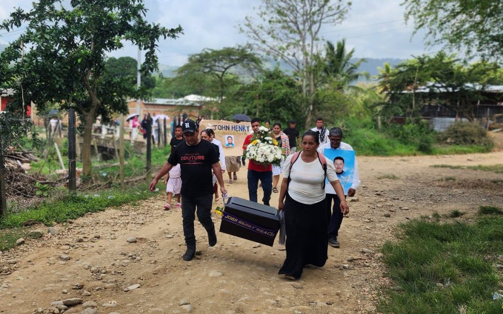 Un grupo de familiares camina en procesión por un camino de tierra. En el centro, un hombre con gorra negra y una mujer con falda larga sostienen conjuntamente un pequeño cofre fúnebre que lleva inscrito el nombre "JHON HENRY GRACIANO". Detrás de ellos, otra persona lleva un arreglo de flores blancas y el hombre con el retrato del joven acompaña el cortejo.