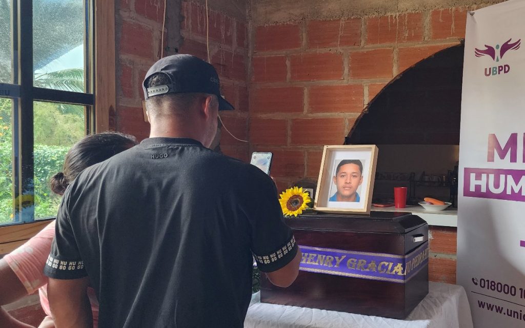 Fotografía desde la parte trasera de un hombre con gorra negra y camiseta del mismo color, quien sostiene un teléfono celular para tomar una foto del altar de entrega digna. Sobre la mesa cubierta con mantel blanco reposa un cofre fúnebre de madera con la cinta con el nombre de Jhon Henry Graciano, junto a un girasol y un marco de madera con la foto del joven desaparecido. Al fondo se divisa el pendón de la UBPD.