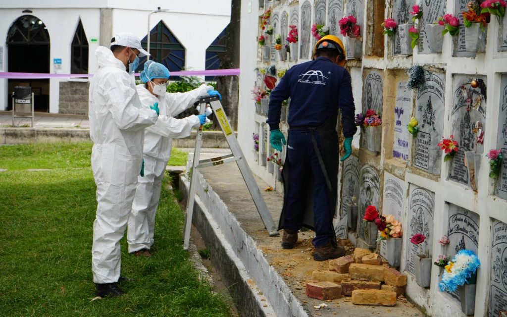 Tres personas del equipo forense con traje de protección blanco y equipo de cementerio trabajan junto a una pared de nichos en una exhumación. Frente a la pared hay ladrillos apilados en el suelo.