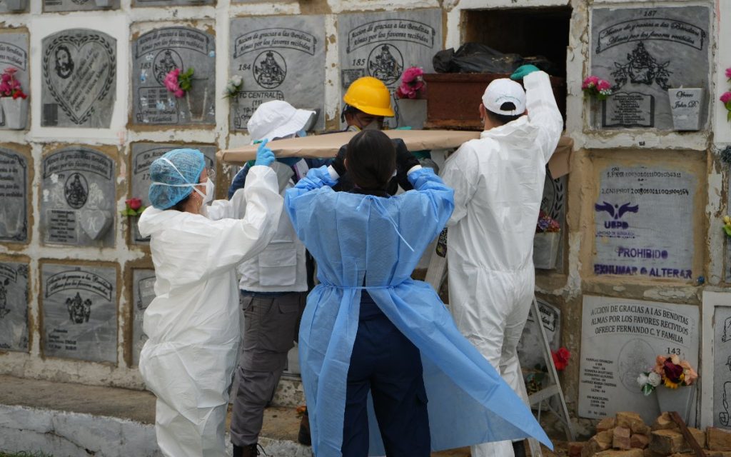 Personal con trajes bioseguros y cascos extraen con cuidado un ataúd de madera de un nicho en un pabellón del cementerio.