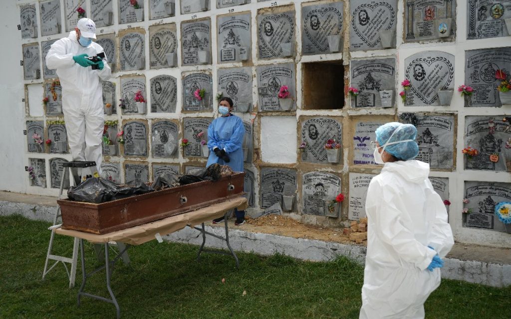 Un fotógrafo forense subido a una escalera toma fotografías al contenido de un ataúd ubicado sobre una mesa plegable, mientras dos profesionales de salud y bioseguridad observan la diligencia frente a los nichos.