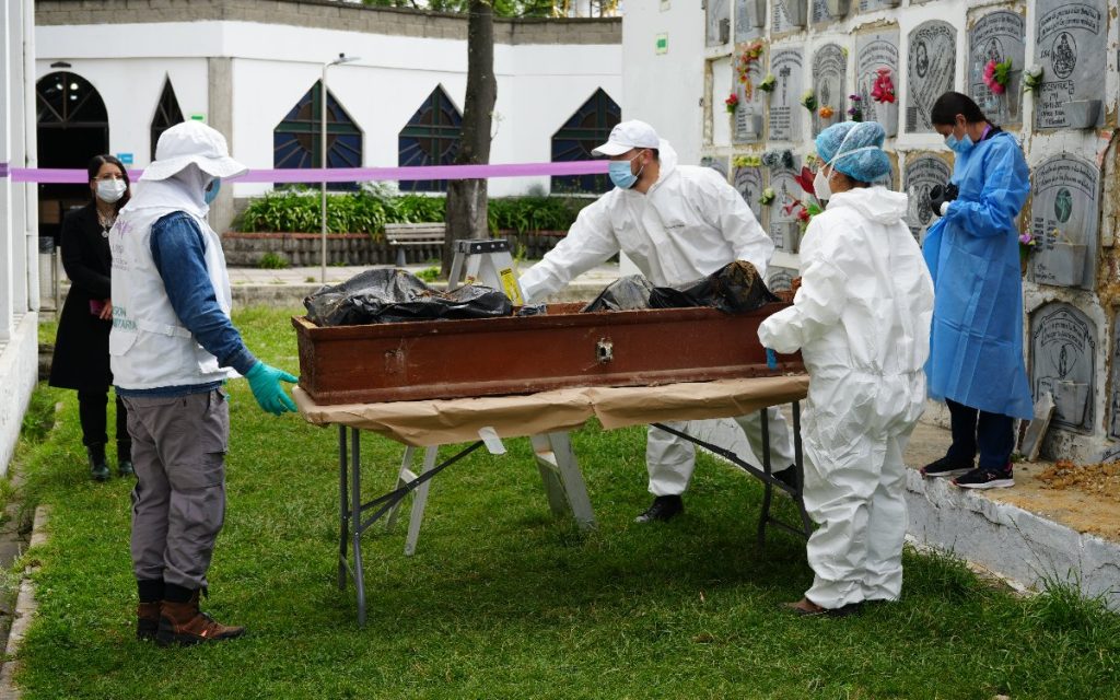 Cuatro profesionales forenses y de búsqueda humanitaria con trajes y batas de protección rodean una mesa con el ataúd exhumado en las áreas verdes del cementerio.