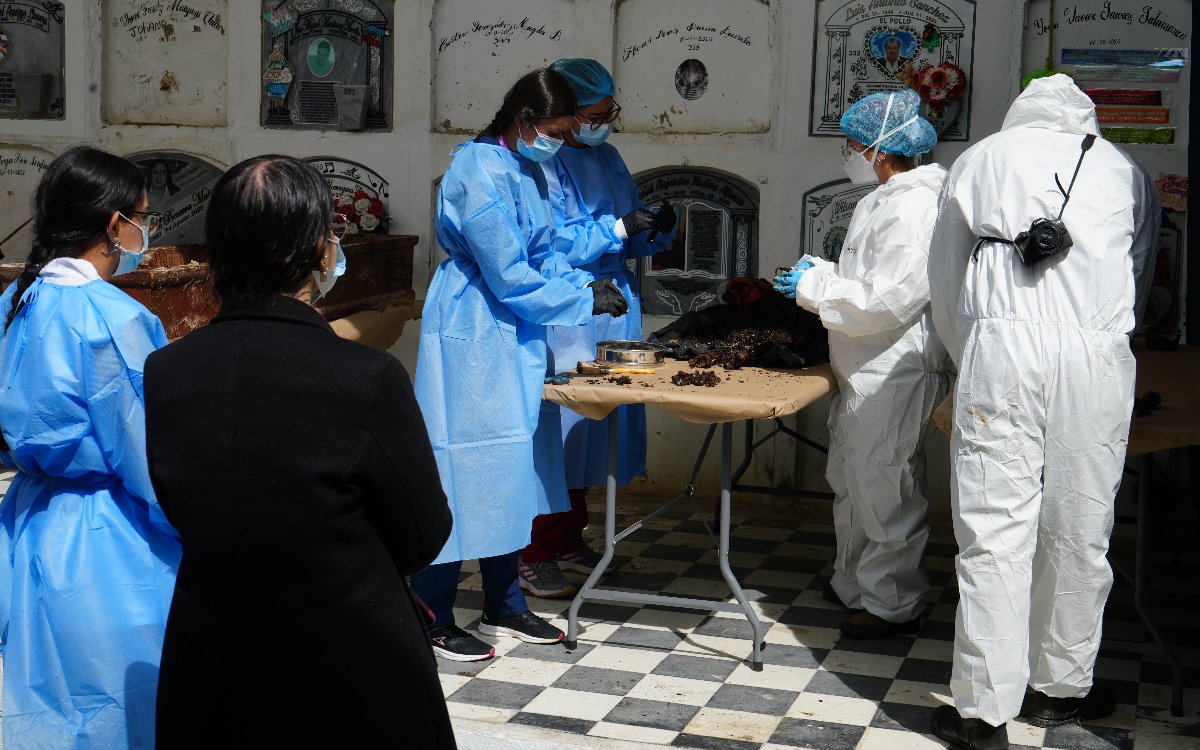 Integrantes del equipo técnico-forense vestidos con batas azules y trajes blancos analizan muestras sobre una mesa, bajo la atenta mirada de observadores en el cementerio.