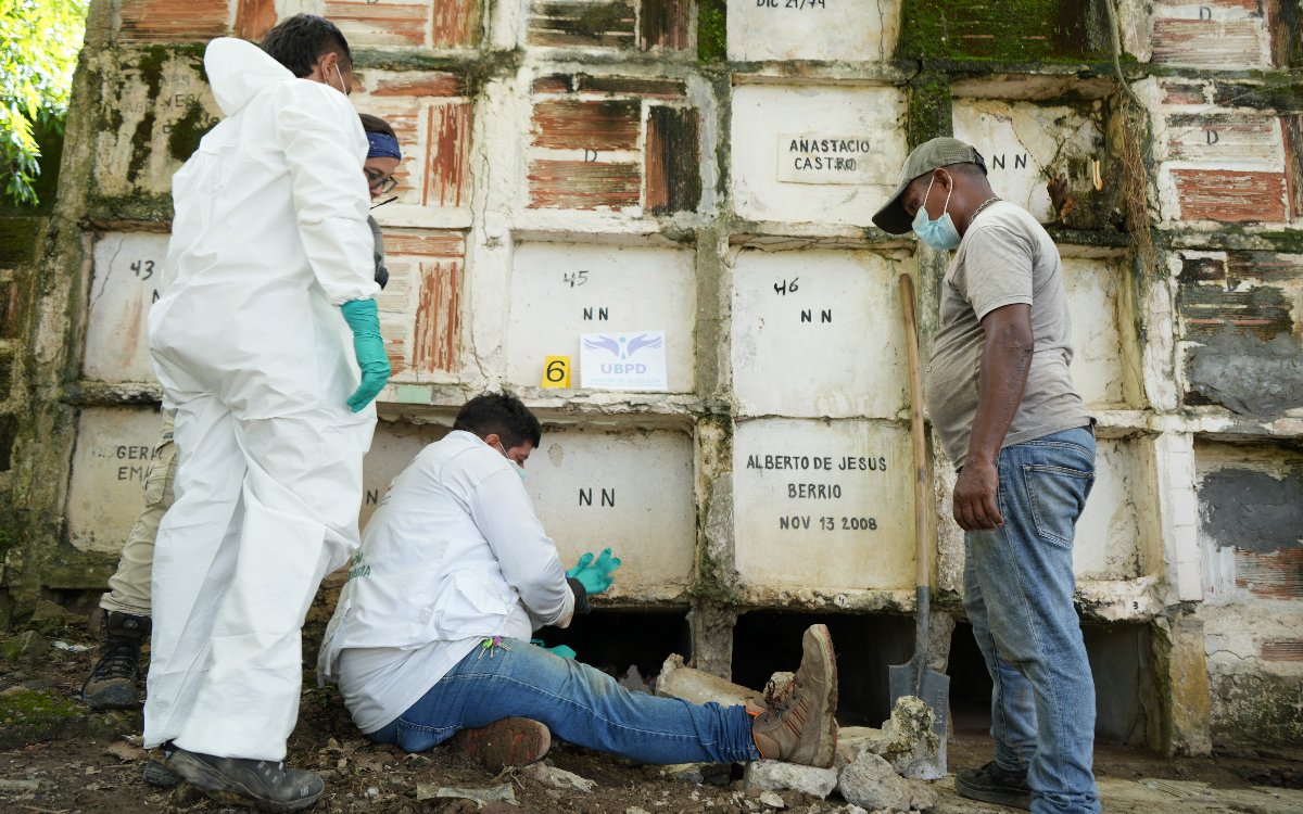 Tres integrantes de un equipo forense de la Unidad de Búsqueda de Personas dadas por Desaparecidas (UBPD) trabajan frente a una pared de bóvedas funerarias de concreto. Dos llevan trajes de protección biológica blancos y guantes verdes; uno de ellos está sentado en el suelo examinando una de las cavidades inferiores. A la derecha, un hombre con mascarilla y pala observa el procedimiento.