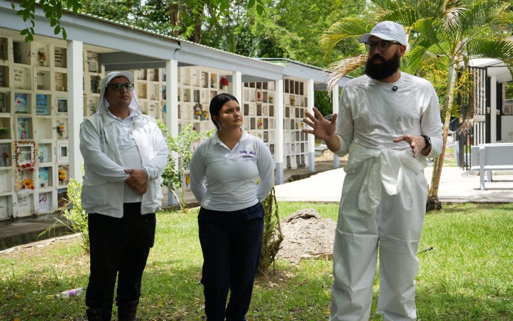 Un hombre con barba, gorra blanca y traje de bioseguridad blanco gesticula con las manos mientras se dirige a dos compañeros de la UBPD (un hombre y una mujer vestida con camiseta institucional blanca), quienes lo escuchan atentos de pie sobre el césped de un cementerio con pabellones de bóvedas al fondo.