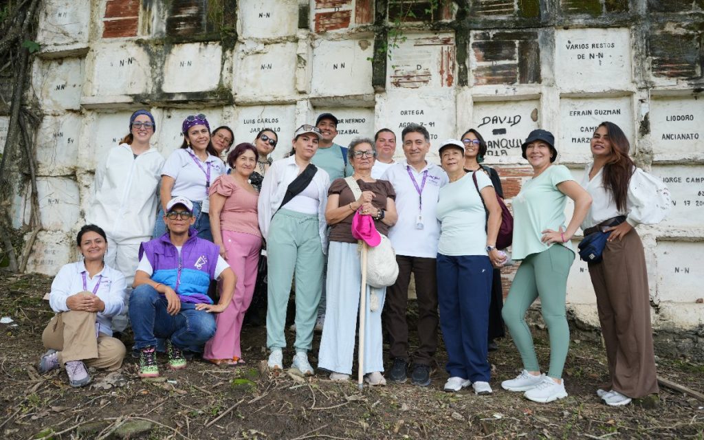 Foto grupal de aproximadamente quince personas, incluyendo miembros del equipo de la UBPD e integrantes de la comunidad o familiares, posando alineados frente a una pared antigua de bóvedas en un cementerio. En el centro, una mujer mayor sostiene un bastón de madera y un sombrero rosa.
