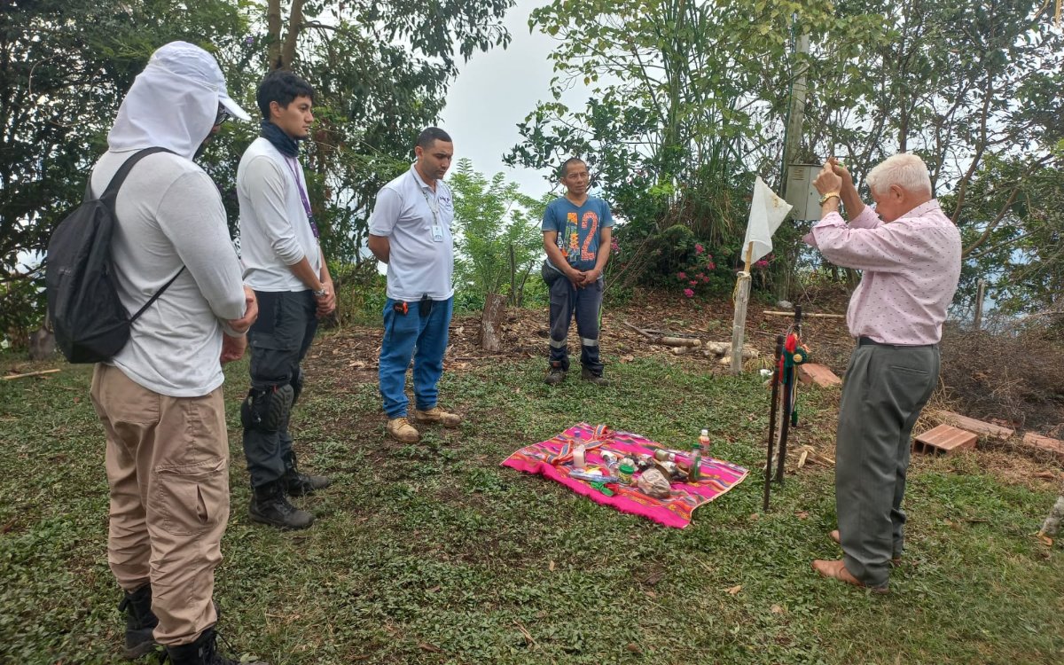 Cinco personas están de pie al aire libre en un espacio verde rodeado de vegetación. Frente a ellas hay una manta colorida extendida en el césped con diversos recipientes y objetos ceremoniales. A la derecha, un hombre mayor con camisa clara alza las manos en actitud de bendición o ritual.