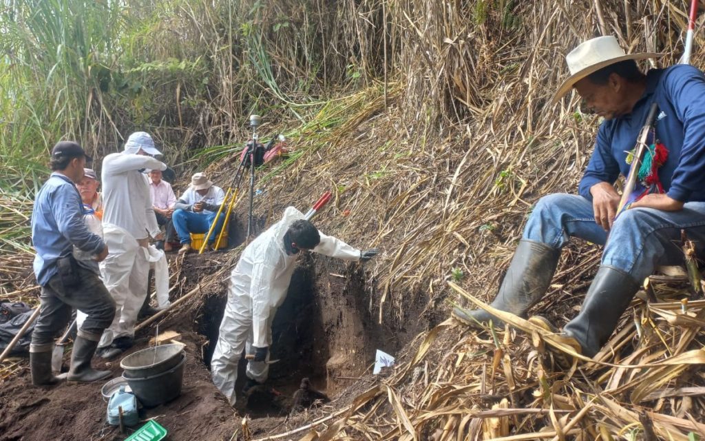 Un hombre vistiendo traje blanco de bioseguridad y otro con morral rojo y jeans avanzan por un camino de maleza seca en una ladera. En la parte inferior derecha se observa un letrero con el logo de la Unidad de Búsqueda de Personas dadas por Desaparecidas (UBPD).