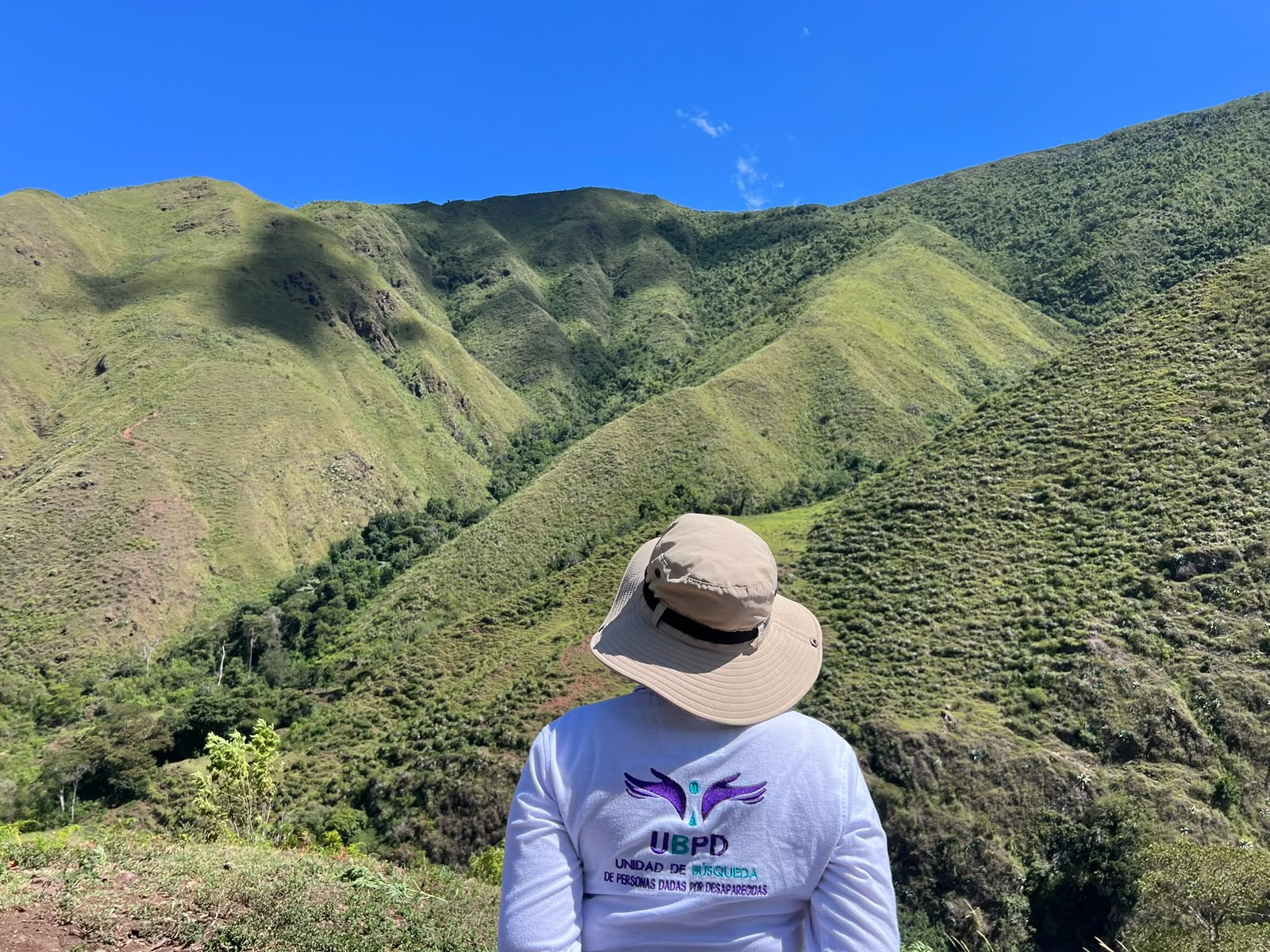 Plano medio desde atrás de una persona de la UBPD portando un sombrero de ala ancha y una camiseta blanca con el logo de la entidad bordado en la espalda. Al frente se aprecia un amplio paisaje montañoso con colinas verdes cubiertas de vegetación, característico de la Serranía del Perijá, bajo un cielo despejado