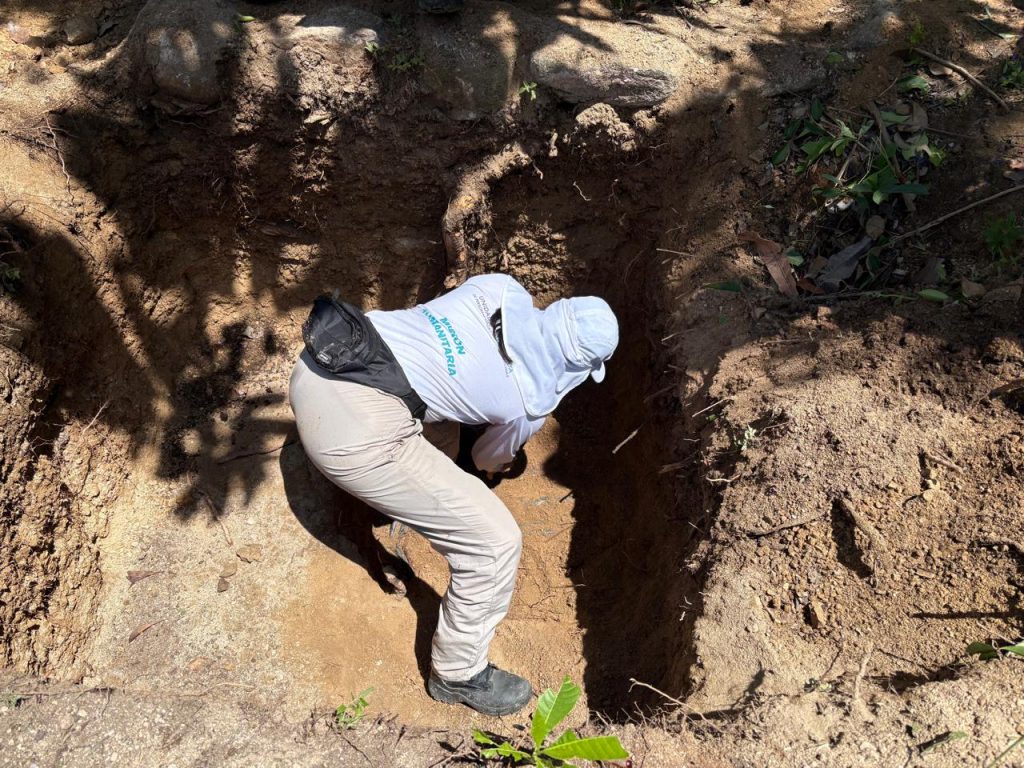 Una persona con uniforme de la UBPD (camisa blanca con la leyenda "Misión Humanitaria", pantalón claro y gorro protector) inclinada de espaldas dentro de una fosa rectangular excavada en la tierra. Se encuentra trabajando minuciosamente al nivel del suelo en un terreno rodeado de sombras y raíces.