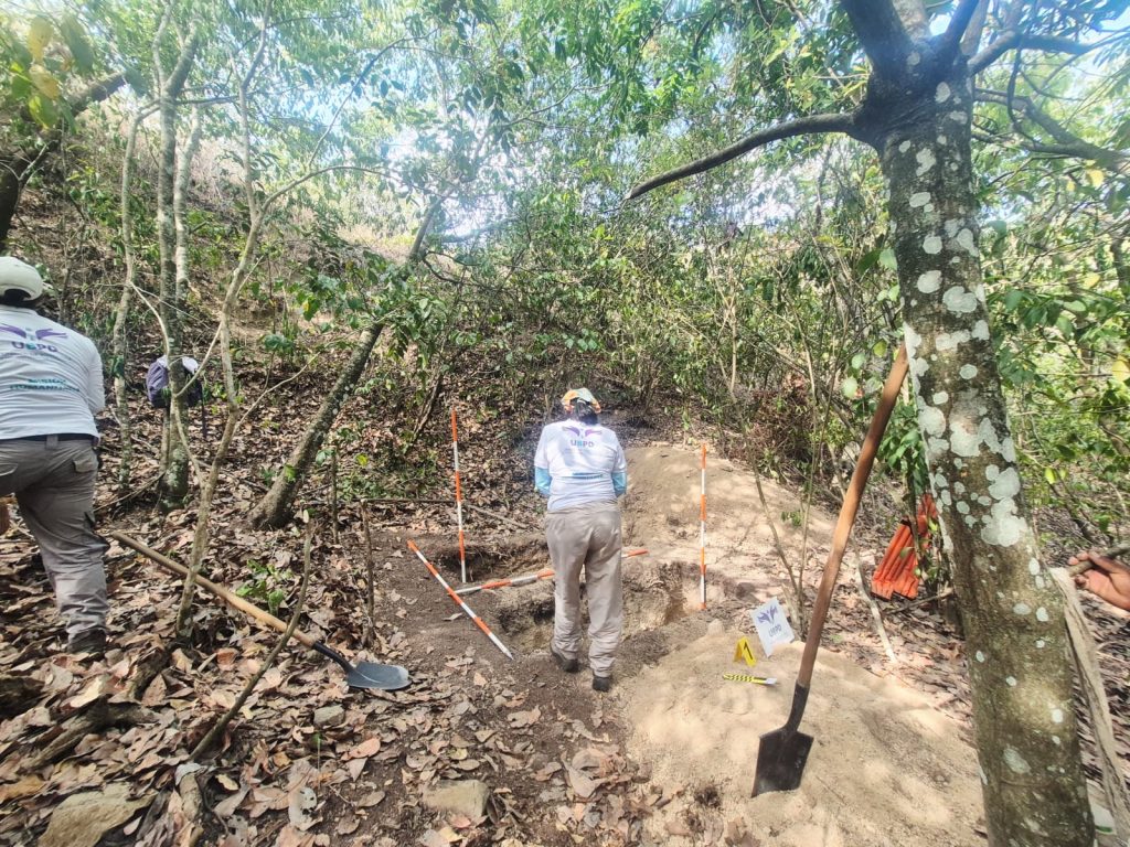 Plano general en una ladera boscosa cubierta de hojas secas y árboles. Dos integrantes de la UBPD realizan labores en un cuadrante de excavación delimitado por vara de medición ortogonales de color naranja y blanco. En el suelo se observan palas, un marcador de evidencia amarillo con el número 1 y una señalización con el logo de la UBPD.