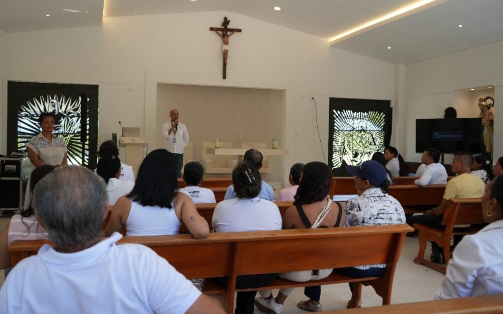Vista desde la parte trasera del interior de una capilla iluminada. En el fondo, un hombre con camisa blanca habla sosteniendo un micrófono ante un auditorio compuesto por hombres y mujeres sentados en bancas de madera. En la pared principal del altar se destaca una gran cruz con la figura de Cristo.
