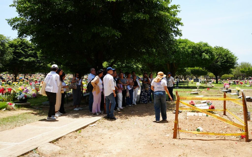 Un grupo de hombres y mujeres permanece de pie bajo la sombra de árboles en un cementerio rodeado de lápidas y flores. Una funcionaria de la Unidad de Búsqueda de Personas dadas por Desaparecidas (UBPD), vestida con chaleco institucional y sombrero, les habla señalando un área de excavación acordonada con cinta amarilla de advertencia.