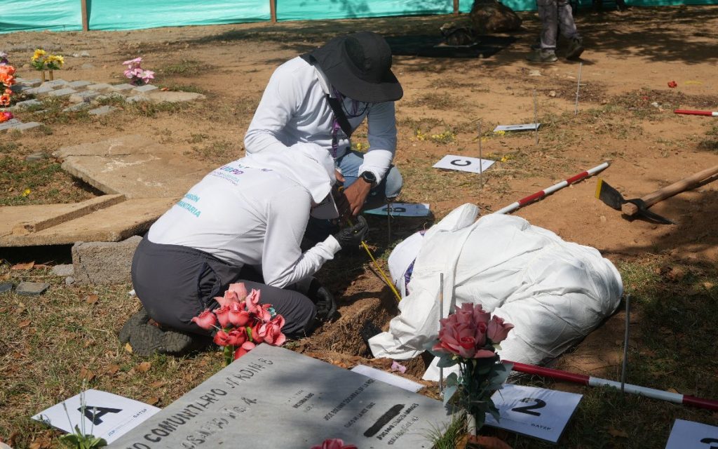 Plano en ángulo picado donde tres profesionales de la UBPD, vistiendo trajes de protección blancos y gorros, trabajan sobre el suelo de tierra en una tumba de cementerio. Usan una cinta métrica y señalizadores alfanuméricos en el terreno cerca de arreglos florales y una lápida de piedra.