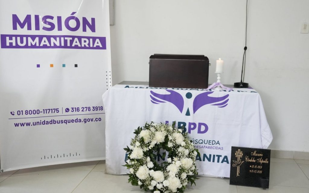 En primer plano se observa una corona fúnebre de flores blancas frente a una mesa ceremonial vestida con una tela que lleva el logo de la Unidad de Búsqueda de Personas dadas por Desaparecidas (UBPD). Sobre la mesa reposa un cofre fúnebre pequeño de madera, una vela blanca sobre un candelabro y al lado derecho una placa conmemorativa negra grabado en dorado con una cruz y el nombre Macario Córdoba Asprilla (12-10-1959 / 10-07-2026). A la izquierda destaca un pendón institucional con los datos de contacto y la frase "Misión Humanitaria".