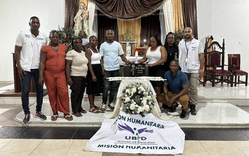 Fotografía grupal tomada al interior de un templo o capilla. Nueve personas (familiares y equipo humanitario de la UBPD vestidas con chalecos blancos) rodean una pequeña mesa ceremonial cubierta con la bandera de Misión Humanitaria, una corona de flores blancas y la placa conmemorativa a nombre de Macario Córdoba Asprilla. Al fondo se observa el altar mayor con la imagen de una virgen.