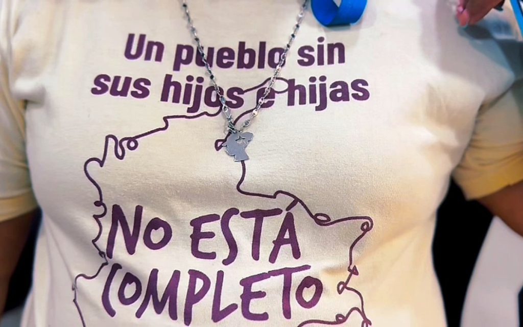 Plano medio del torso de una persona vistiendo una camiseta de color crema o blanco hueso. La prenda tiene una ilustración en líneas moradas que bordea el texto y dice en letras mayúsculas y minúsculas: «Un pueblo sin sus hijos e hijas NO ESTÁ COMPLETO». La persona lleva además un collar con un dije metálico al cuello.