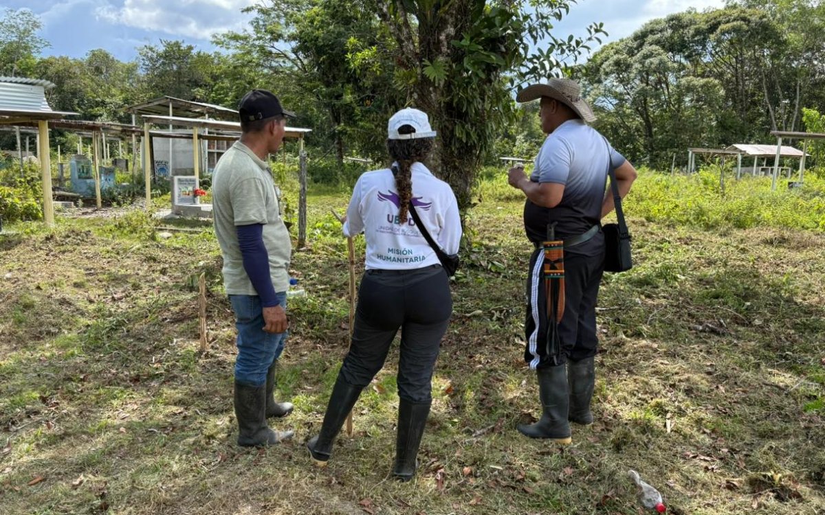 Tres personas permanecen de pie de espaldas a la cámara conversando en un campo de pasto recortado en zona rural. La persona del centro lleva gorra blanca y una camiseta blanca con el logotipo y texto visible de «UBPD - MISIÓN HUMANITARIA». A sus costados hay dos hombres con botas de caucho y sombreros o gorras. De fondo se aprecia vegetación alta y pequeñas estructuras rústicas.