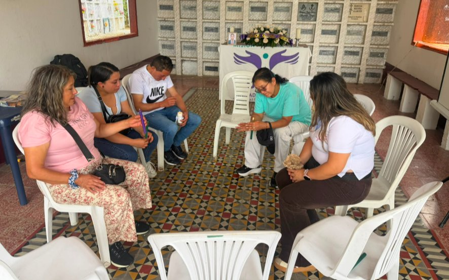 Imagen en plano cenital de un grupo de hombres y mujeres sentados en sillas plásticas blancas dispuestas en círculo. Varias de las personas sostienen velas encendidas en sus manos en un ambiente reflexivo y conmemorativo. Al fondo, frente a una pared de nichos funerarios, hay una mesa cubierta con un mantel blanco con el logotipo de la UBPD («Unidad de Búsqueda de Personas dadas por Desaparecidas») y un arreglo floral.