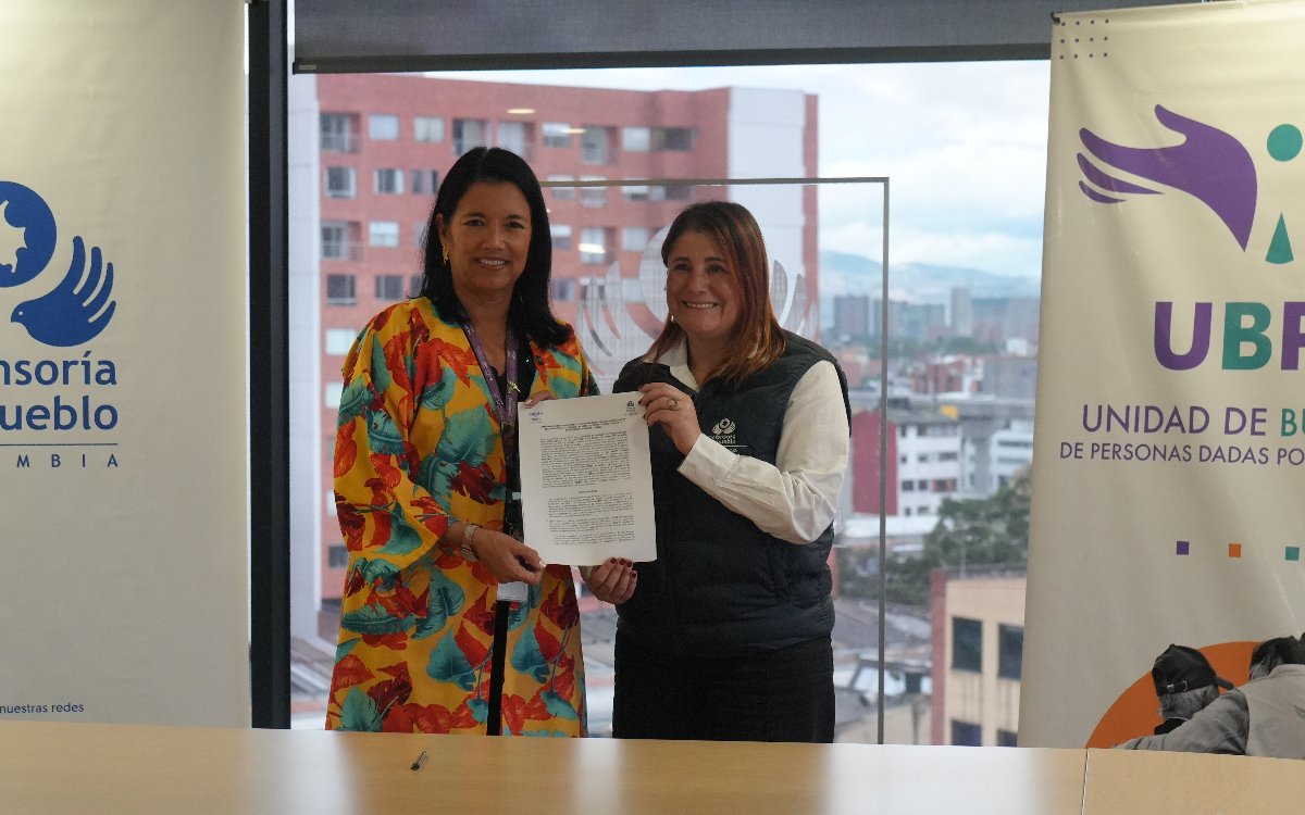 Fotografía en plano medio de la firma del memorando de entendimiento entre la Defensoría del Pueblo y la UBPD. Dos representantes sostienen el documento firmado en el centro de la escena, sonriendo hacia la cámara. Al fondo se observa un ventanall con vista a la ciudad y dos pendones de marca a los lados con los logotipos de la Defensoría del Pueblo de Colombia y la Unidad de Búsqueda de Personas dadas por Desaparecidas (UBPD).