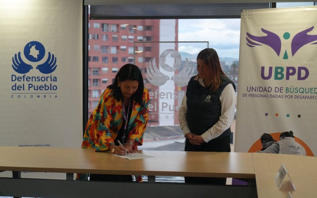 Fotografía de la firma del convenio. La representante con saco amarillo floral aparece inclinada sobre una gran mesa de madera firmando el memorando con un bolígrafo. A su derecha, la funcionaria de la Defensoría del Pueblo permanece de pie observando el acto con las manos cruzadas. A los lados se aprecian los pendones con la imagen corporativa de ambas instituciones.