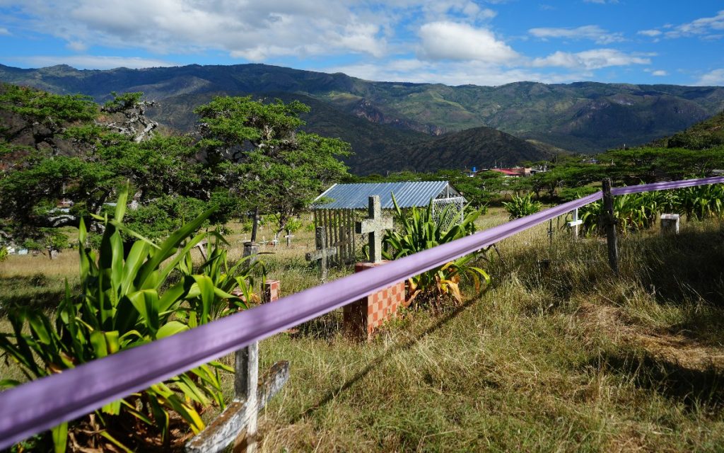 Fotografía panorámica de un cementerio rodeado de vegetación y campos secos, con montañas de fondo bajo un cielo parcialmente nublado. En primer plano, una cinta de delimitación color morado atraviesa la escena. Hay tumbas con cruces de piedra sobre bases de ladrillo y plantas verdes a los lados.