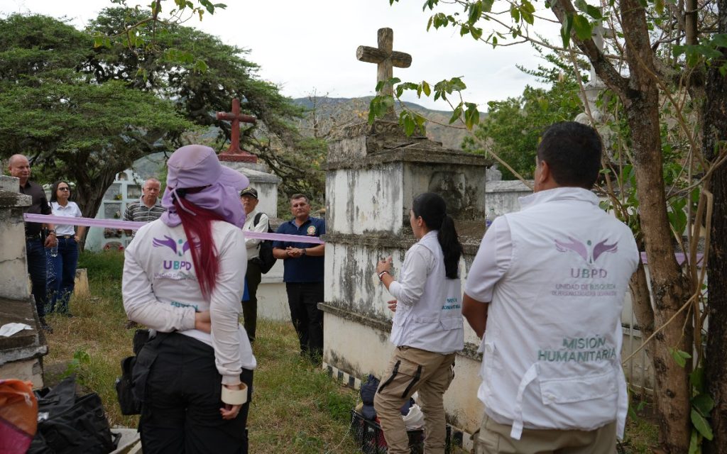 Grupo de personas reunidas junto a un mausoleo de piedra coronado con una cruz. En primer plano, dos funcionarias de la UBPD llevan chalecos blancos con el logo institucional y la leyenda "Misión Humanitaria". Al fondo, campesinos y miembros de la comunidad escuchan atentos detrás de una cinta de delimitación morada.