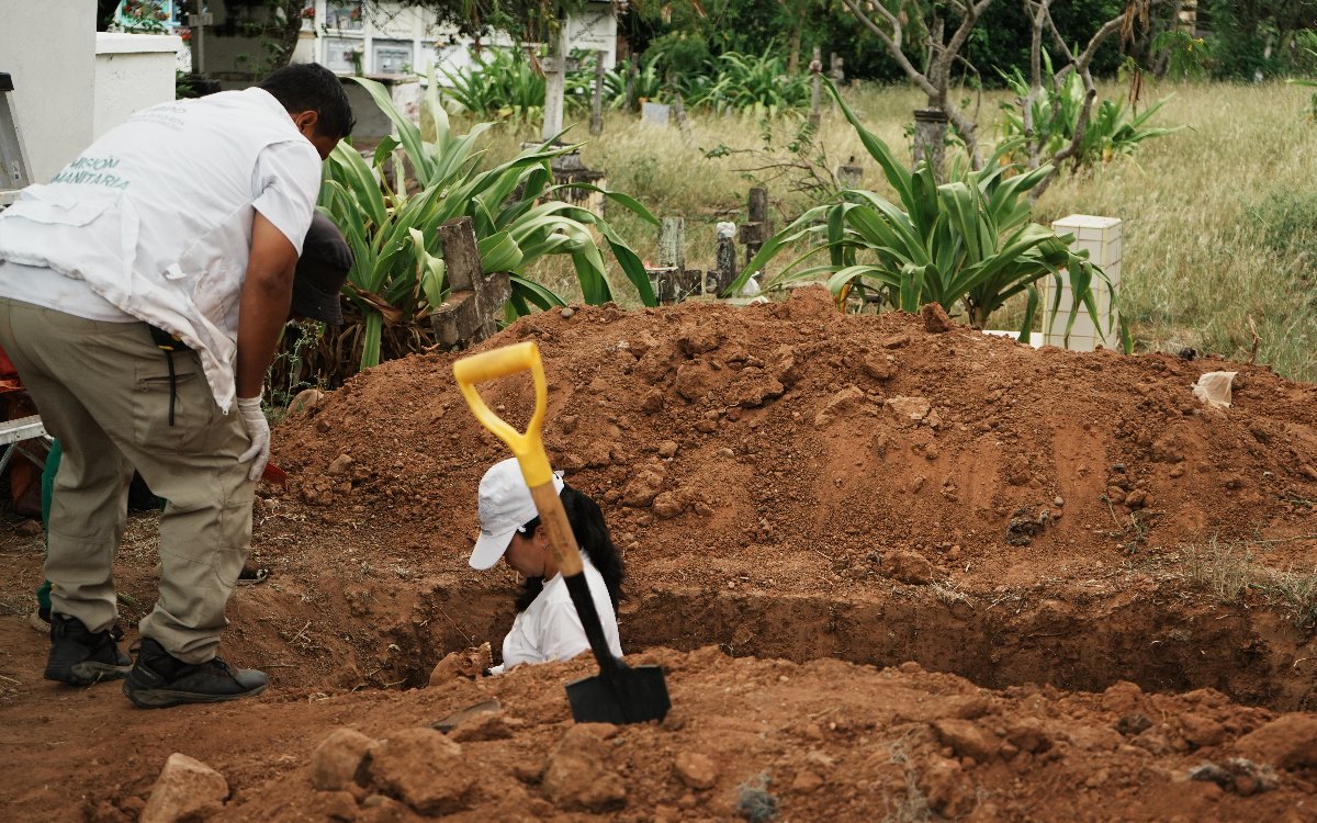 Un equipo de prospección forense realiza labores de excavación. Una mujer con gorra blanca y camiseta del mismo color está inclinada en el interior de una fosa rodeada por un cúmulo de tierra marrón. A su lado hay una pala clavada en la tierra y un compañero inclinado observando el proceso desde el borde.