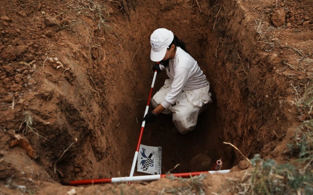 Fotografía desde un ángulo superior que muestra a una profesional de la UBPD vestida de blanco, con gorra y guantes negros, dentro de una fosa rectangular excavada en la tierra. Utiliza reglas métricas de color rojo y blanco para acotar el hallazgo y posiciona una etiqueta de registro con el logo de la UBPD.