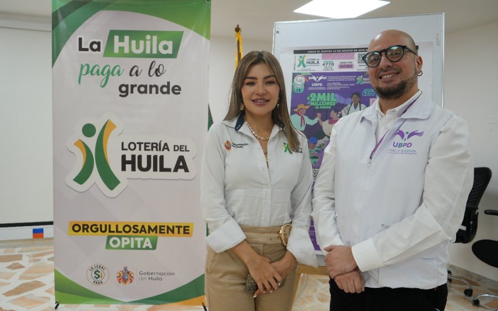 Fotografía en interiores de dos personas de pie lado a lado. A la izquierda, una mujer viste blusa blanca de la Gobernación del Huila y pantalón beis. A la derecha, un hombre con gafas y chaleco blanco de la Unidad de Búsqueda de Personas dadas por Desaparecidas (UBPD). Detrás de ellos hay un pendón publicitario con la frase "La Huila paga a lo grande" y un tablero con una ampliación del billete de lotería del 25 de agosto de 2026.