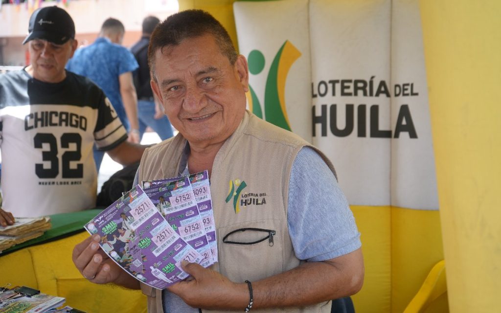 Plano medio de un vendedor de lotería maduro luciendo un chaleco beis con el logo de la Lotería del Huila. El vendedor sonríe a la cámara mientras sostiene en sus manos varios billetes morados del sorteo de la Lotería del Huila alusivos a la búsqueda de personas desaparecidas.