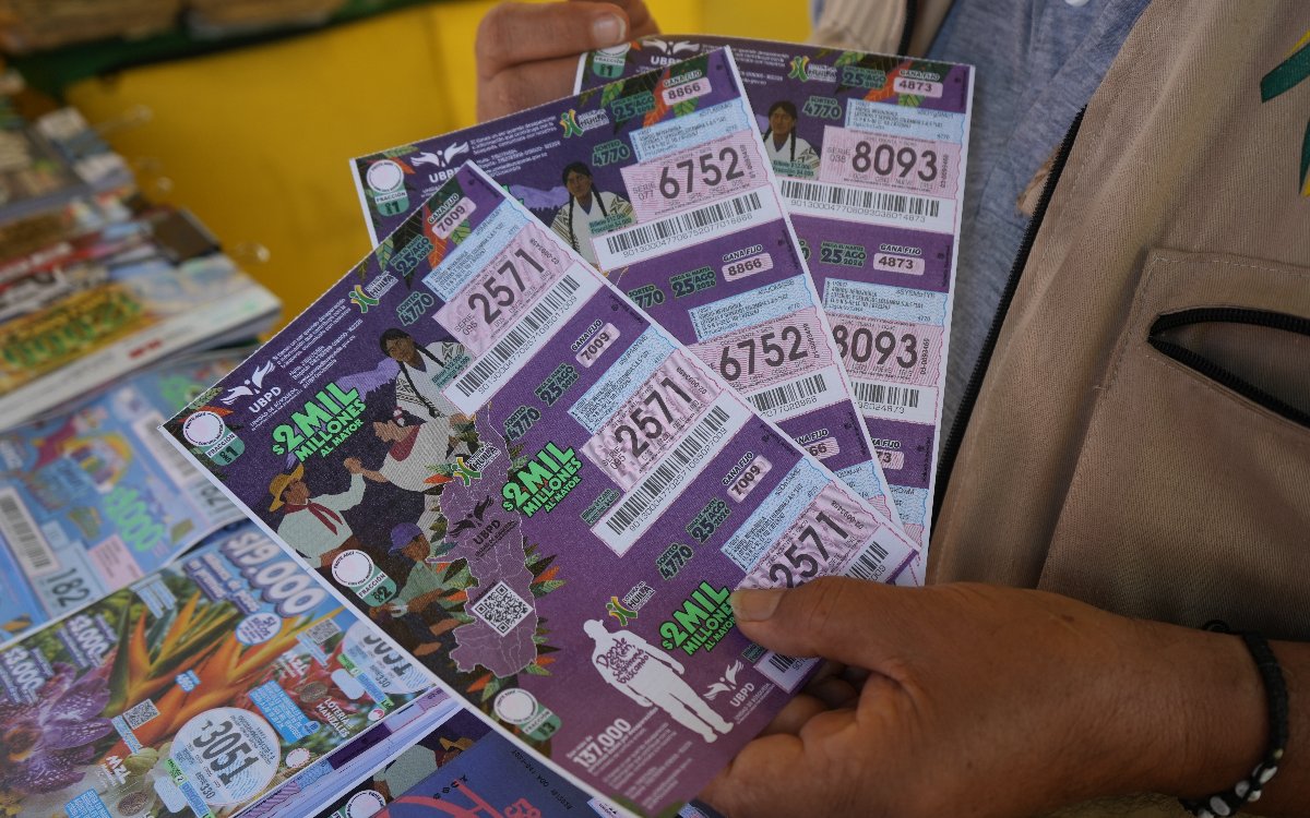 Detalle en primer plano enfocado en los billetes de color morado de la Lotería del Huila. Se observa claramente el diseño del billete con el premio mayor de 2.000 millones de pesos, el logotipo de la UBPD (Unidad de Búsqueda de Personas dadas por Desaparecidas) e ilustraciones de campesinos e indígenas sobre el mapa de Colombia.